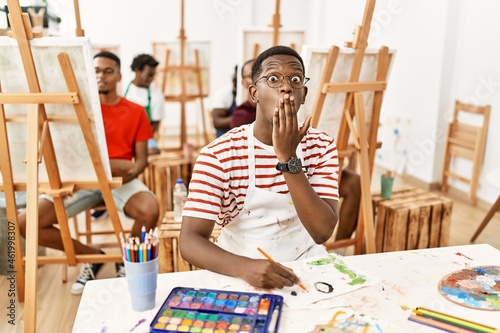 Young african man at art studio covering mouth with hand, shocked and afraid for mistake. surprised expression