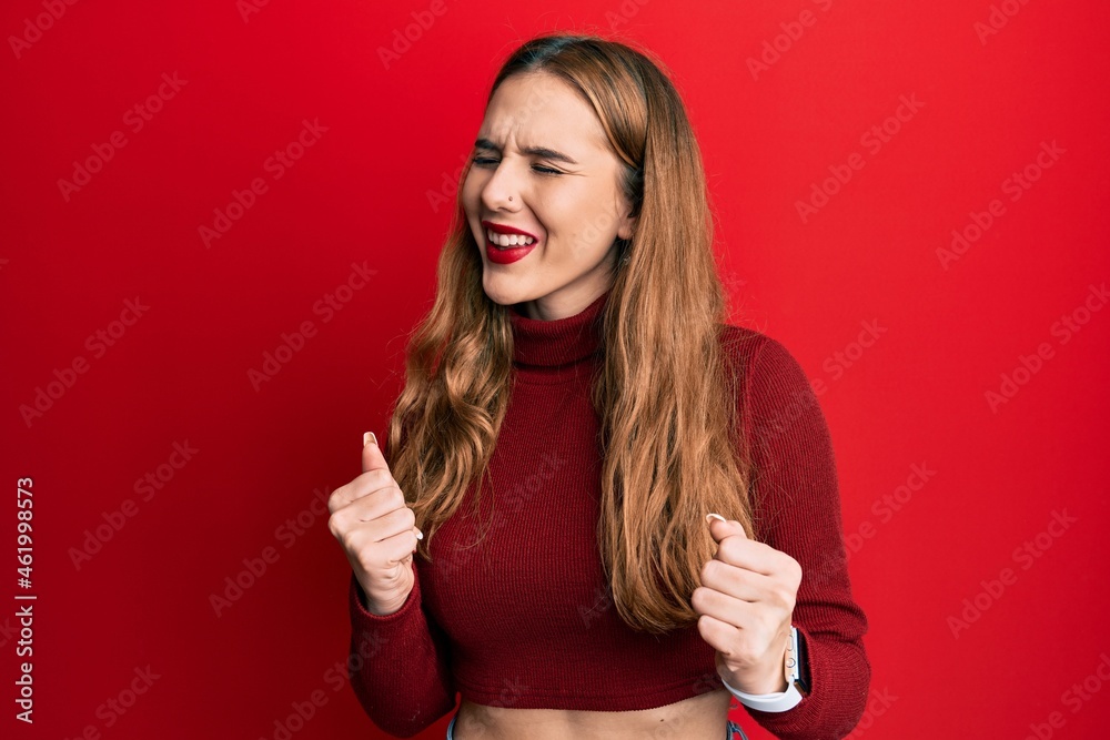 Young blonde woman wearing turtleneck sweater excited for success with arms raised and eyes closed celebrating victory smiling. winner concept. Young blonde woman wearing turtleneck sweater excited for success with arms raised and eyes closed celebrating victory smiling. winner concept.