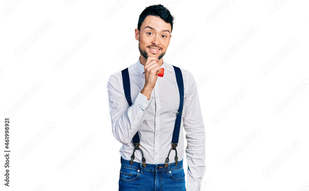 Fototapeta premium Hispanic man with beard wearing hipster look with bow tie and suspenders looking confident at the camera with smile with crossed arms and hand raised on chin. thinking positive.