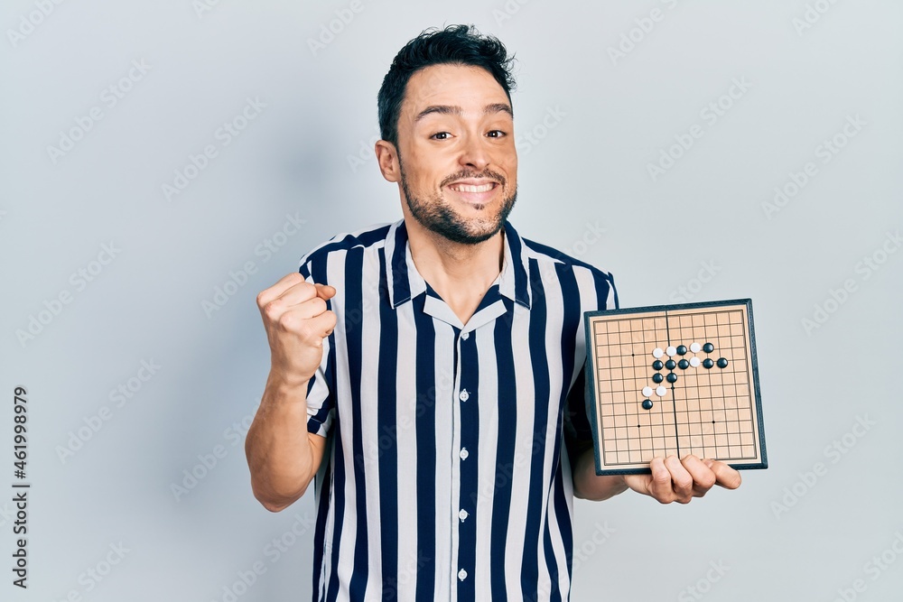 Young hispanic man holding asian go game board screaming proud, celebrating victory and success very excited with raised arm Young hispanic man holding asian go game board screaming proud, celebrating victory and success very excited with raised arm