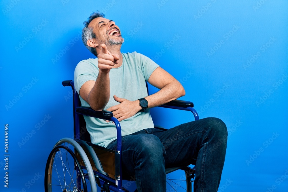 Fototapeta premium Handsome middle age man with grey hair sitting on wheelchair laughing at you, pointing finger to the camera with hand over body, shame expression
