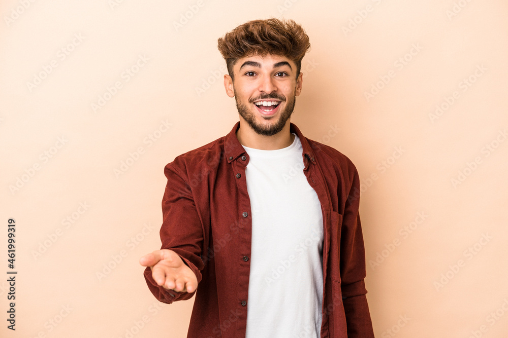 Young arab man isolated on beige background stretching hand at camera in greeting gesture. Young arab man isolated on beige background stretching hand at camera in greeting gesture.