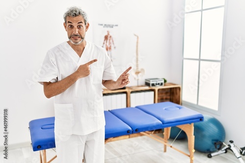 Middle age hispanic therapist man working at pain recovery clinic pointing aside worried and nervous with both hands, concerned and surprised expression