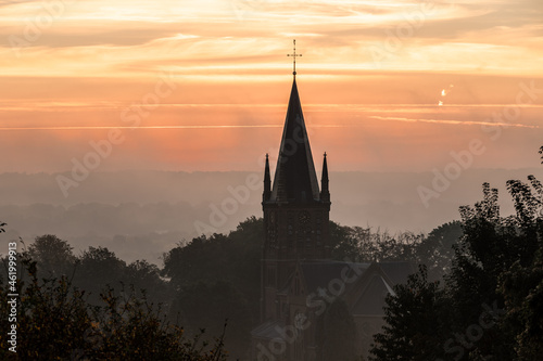 A typical colorful Autumn sunrise in Maastricht with the landscape covered with a layer of fog, leaving only silhouettes visible in the distance, like this tower of a church on the hillside. © KimWillems