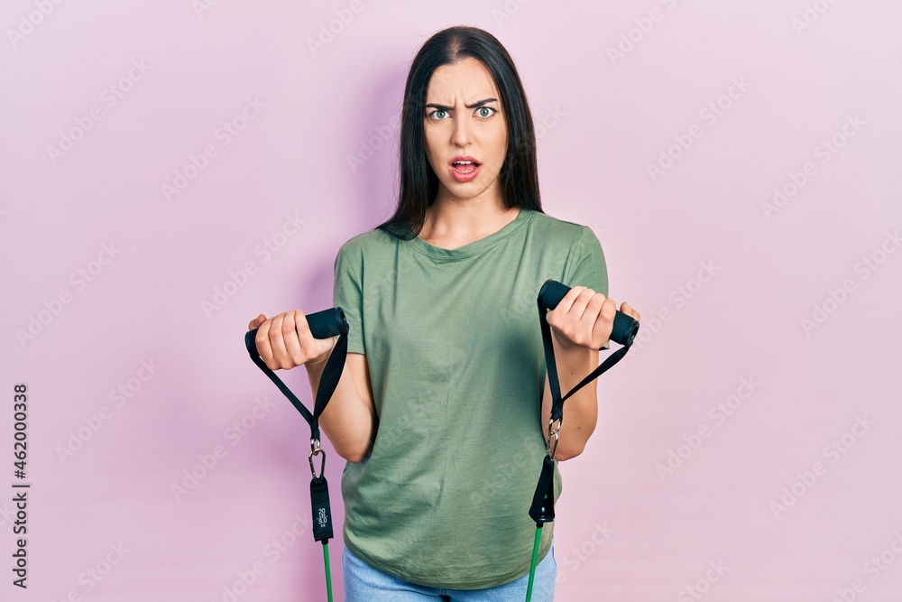 Beautiful woman with blue eyes training arm resistance with elastic arm bands in shock face, looking skeptical and sarcastic, surprised with open mouth
