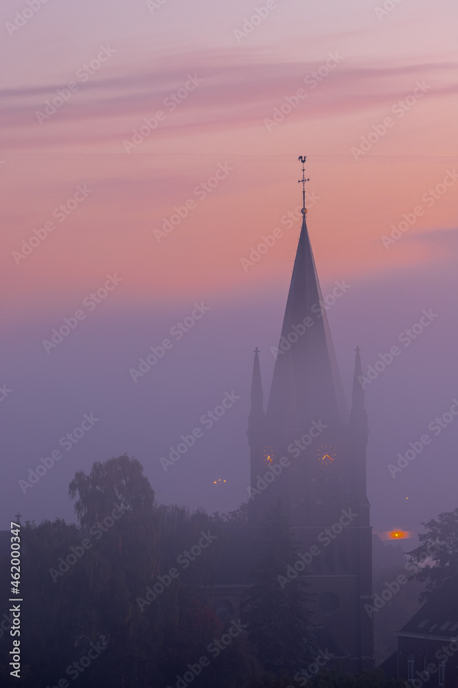 A typical colorful Autumn sunrise in Maastricht with the landscape covered with a layer of fog, leaving only silhouettes visible in the distance, like this tower of a church on the hillside.