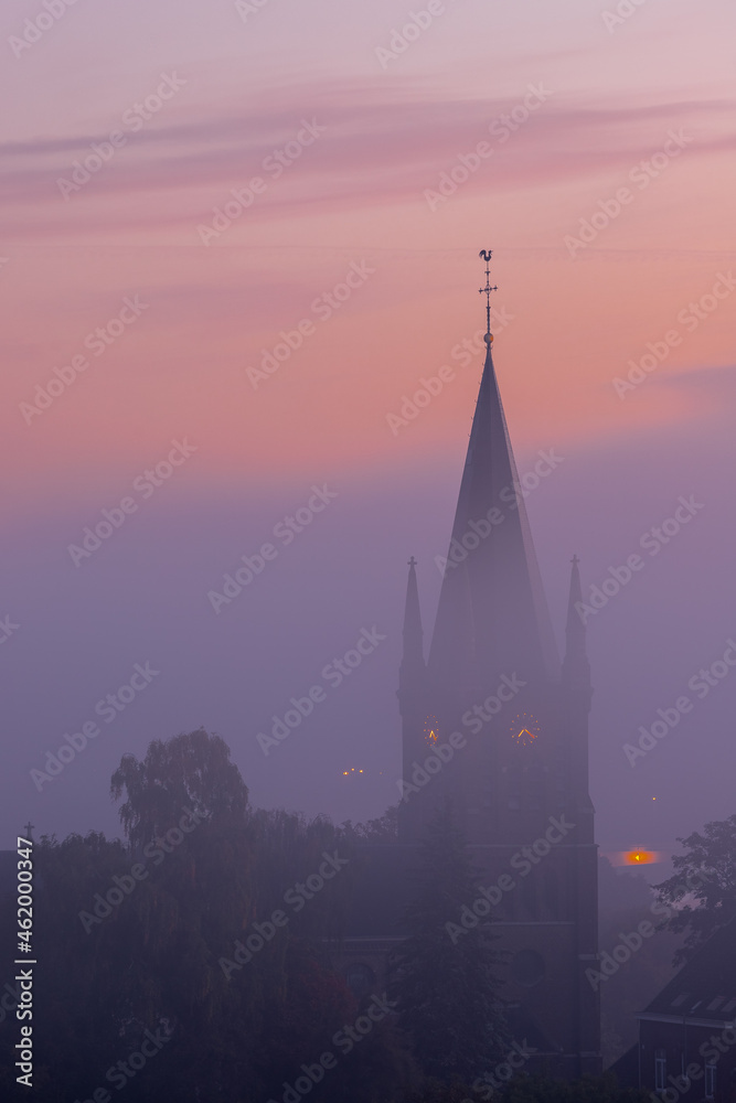 Fototapeta premium A typical colorful Autumn sunrise in Maastricht with the landscape covered with a layer of fog, leaving only silhouettes visible in the distance, like this tower of a church on the hillside.