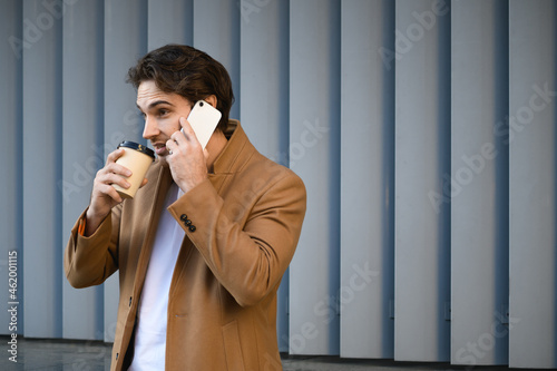 Businessman in coat drinking coffee in paper cup and talking on cellphone outdoors 