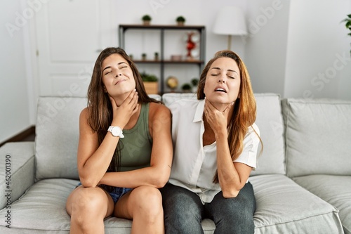 Mother and daughter together sitting on the sofa at home touching painful neck, sore throat for flu, clod and infection