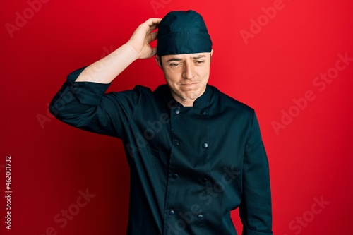 Handsome young man wearing professional cook uniform and hat confuse and wonder about question. uncertain with doubt, thinking with hand on head. pensive concept.