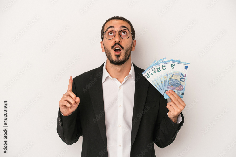 Young caucasian business man holding banknotes isolated on white background pointing upside with opened mouth. Young caucasian business man holding banknotes isolated on white background pointing upside with opened mouth.