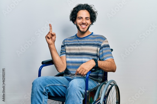 Handsome hispanic man sitting on wheelchair showing and pointing up with finger number one while smiling confident and happy.