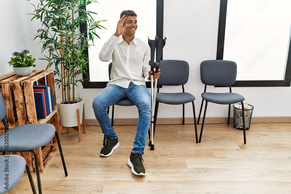 Young hispanic man waiting wearing crutches at waiting room