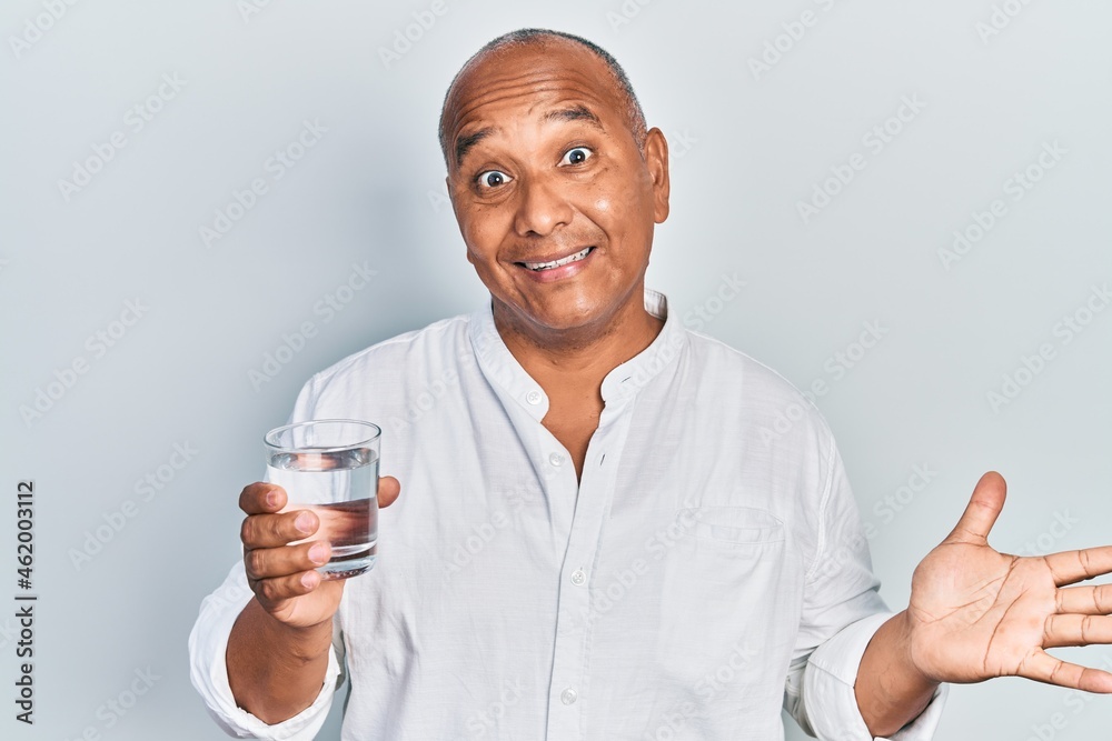 Middle age latin man drinking glass of water celebrating achievement with happy smile and winner expression with raised hand Middle age latin man drinking glass of water celebrating achievement with happy smile and winner expression with raised hand