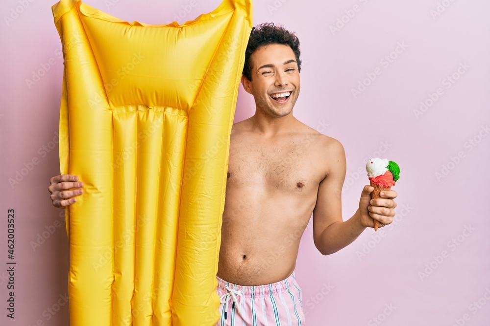 Obraz premium Young handsome man wearing swimsuit holding summer mattress float and icecream winking looking at the camera with sexy expression, cheerful and happy face.
