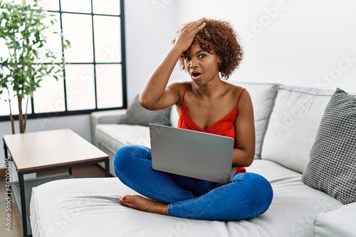 Young african american woman sitting on the sofa at home using laptop surprised with hand on head for mistake, remember error. forgot, bad memory concept.
