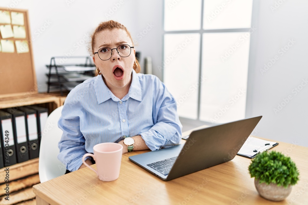 Young redhead woman working at the office using computer laptop in shock face, looking skeptical and sarcastic, surprised with open mouth