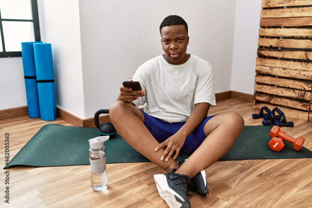 Young african man sitting on training mat at the gym using smartphone looking sleepy and tired, exhausted for fatigue and hangover, lazy eyes in the morning.