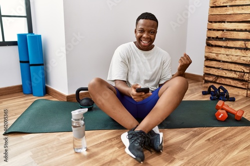 Young african man sitting on training mat at the gym using smartphone very happy and excited doing winner gesture with arms raised, smiling and screaming for success. celebration concept.