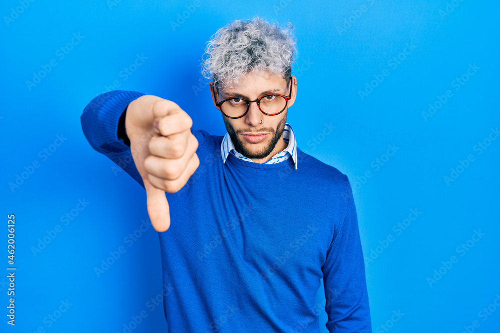 Young hispanic man with modern dyed hair wearing sweater and glasses looking unhappy and angry showing rejection and negative with thumbs down gesture. bad expression. Young hispanic man with modern dyed hair wearing sweater and glasses looking unhappy and angry showing rejection and negative with thumbs down gesture. bad expression.