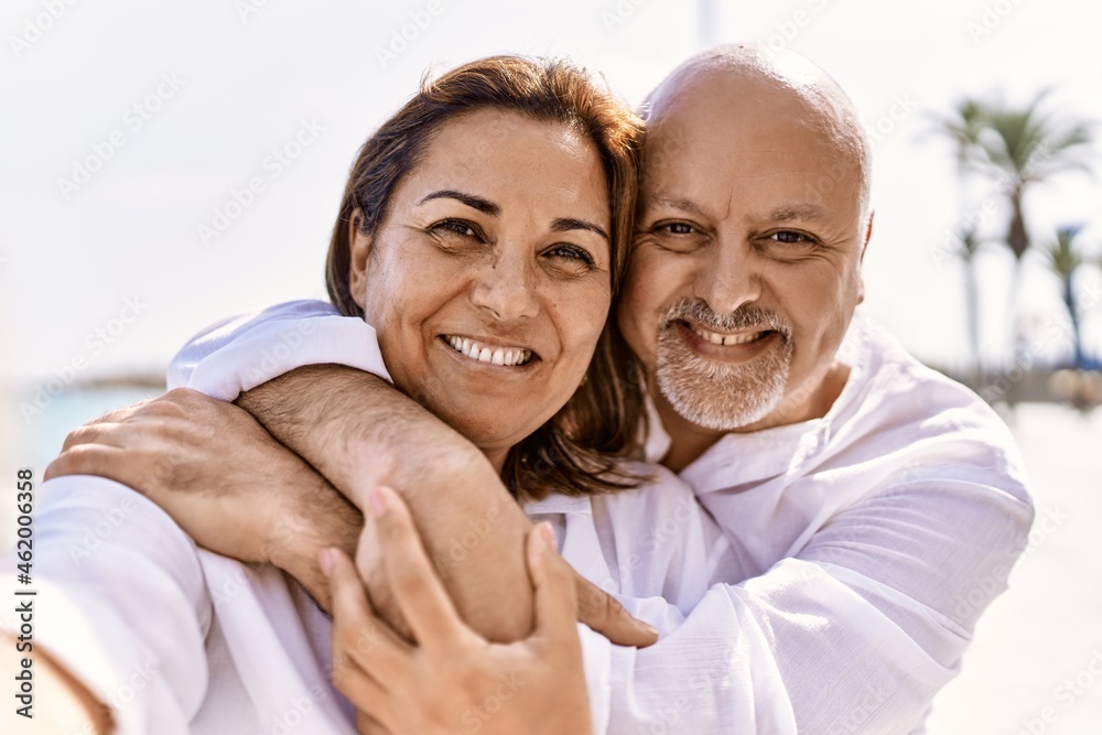Middle age hispanic couple of husband and wife together walking by the beach on a sunny day. Hugging in love on vacation to the seaside taking a selfie picture Middle age hispanic couple of husband and wife together walking by the beach on a sunny day. Hugging in love on vacation to the seaside taking a selfie picture