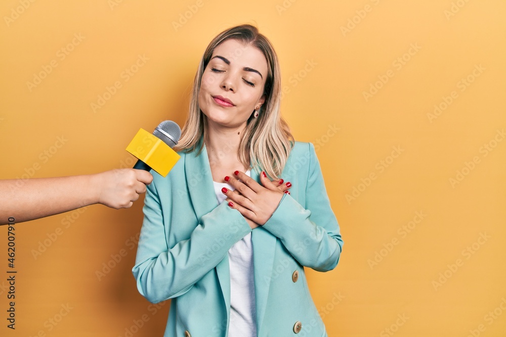 Beautiful caucasian woman being interviewed by reporter holding microphone smiling with hands on chest with closed eyes and grateful gesture on face. health concept. Beautiful caucasian woman being interviewed by reporter holding microphone smiling with hands on chest with closed eyes and grateful gesture on face. health concept.