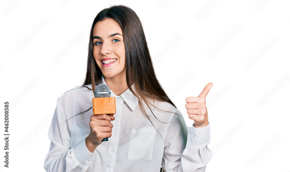 Young brunette teenager holding reporter microphone smiling happy and positive, thumb up doing excellent and approval sign Young brunette teenager holding reporter microphone smiling happy and positive, thumb up doing excellent and approval sign