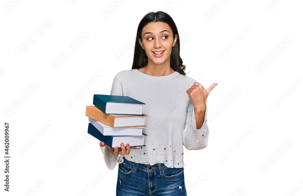 Young hispanic girl holding a pile of books pointing thumb up to the side smiling happy with open mouth Young hispanic girl holding a pile of books pointing thumb up to the side smiling happy with open mouth