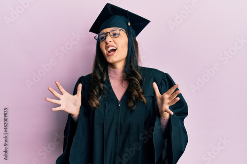 Young hispanic woman wearing graduation cap and ceremony robe crazy and mad shouting and yelling with aggressive expression and arms raised. frustration concept.