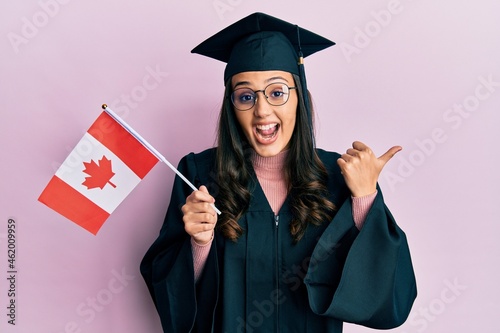 Young hispanic woman wearing graduation uniform holding canada flag pointing thumb up to the side smiling happy with open mouth