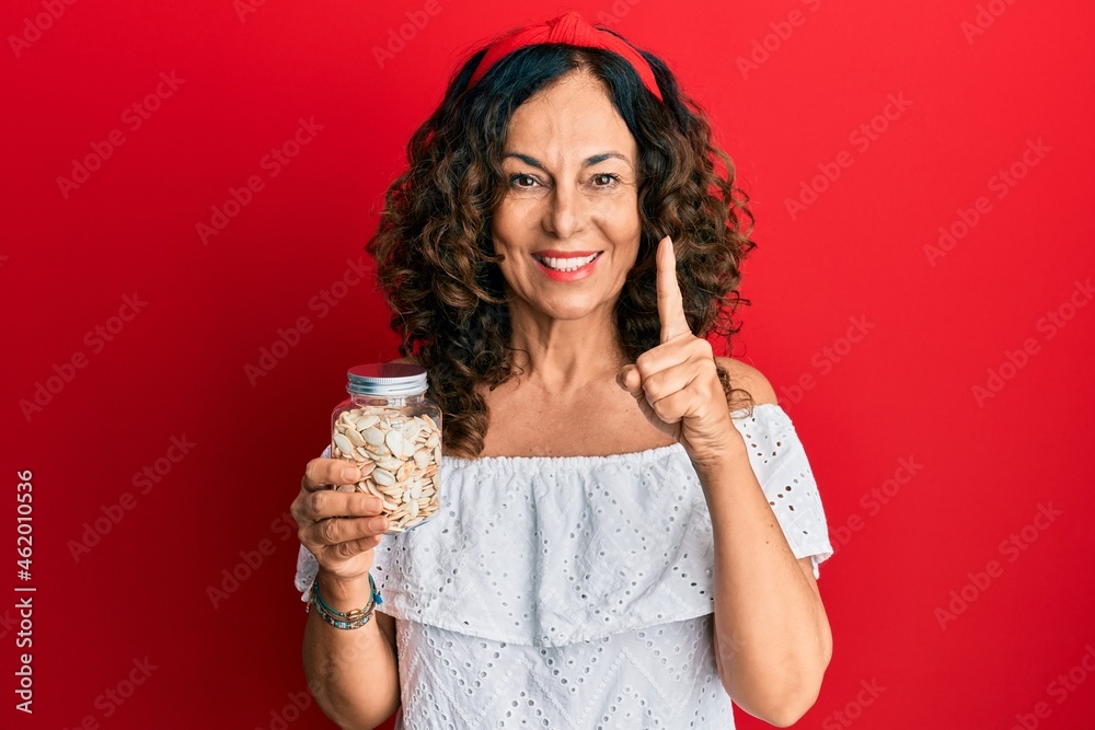 Middle age hispanic woman holding jar with pumpkin seeds smiling with an idea or question pointing finger with happy face, number one