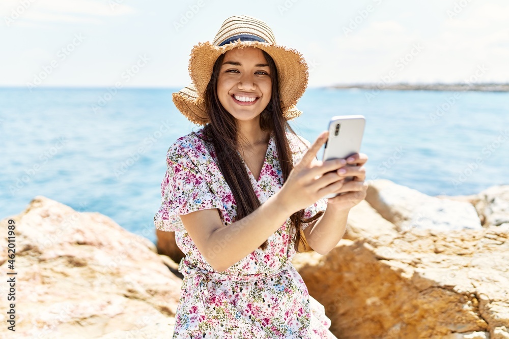 Young latin girl wearing summer hat using smartphone at the beach.