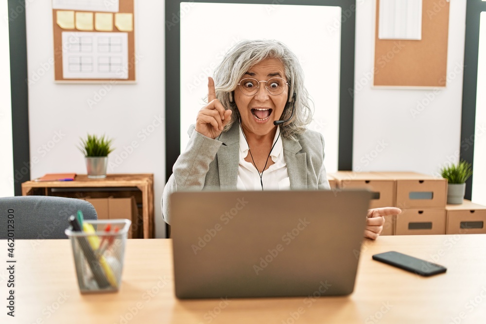 Middle age businesswoman sitting on desk working using laptop at office pointing finger up with successful idea. exited and happy. number one. Middle age businesswoman sitting on desk working using laptop at office pointing finger up with successful idea. exited and happy. number one.