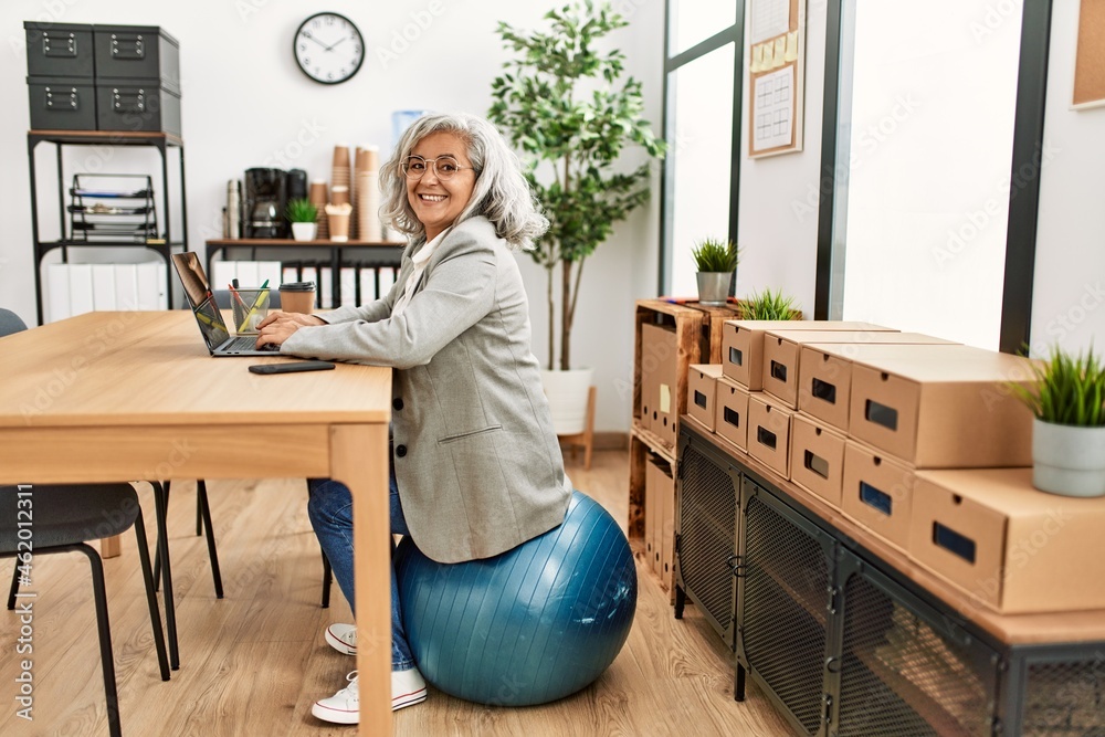 Middle age grey-haired businesswoman sitting on fit ball working at the office. Middle age grey-haired businesswoman sitting on fit ball working at the office.