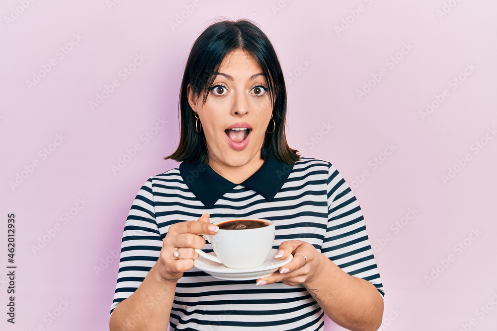 Young hispanic woman drinking a cup of coffee in shock face, looking skeptical and sarcastic, surprised with open mouth Young hispanic woman drinking a cup of coffee in shock face, looking skeptical and sarcastic, surprised with open mouth