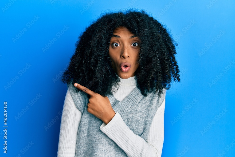 African american woman with afro hair wearing casual winter sweater surprised pointing with finger to the side, open mouth amazed expression. African american woman with afro hair wearing casual winter sweater surprised pointing with finger to the side, open mouth amazed expression.