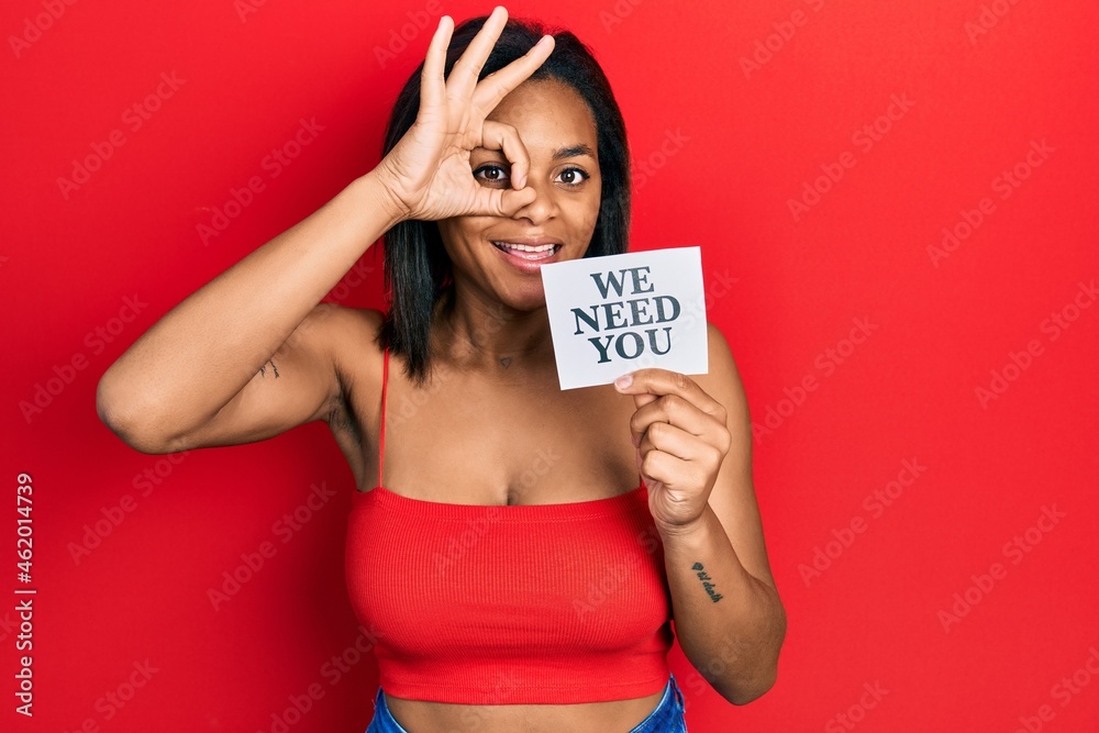 Young african american girl holding we need you paper smiling happy doing ok sign with hand on eye looking through fingers Young african american girl holding we need you paper smiling happy doing ok sign with hand on eye looking through fingers