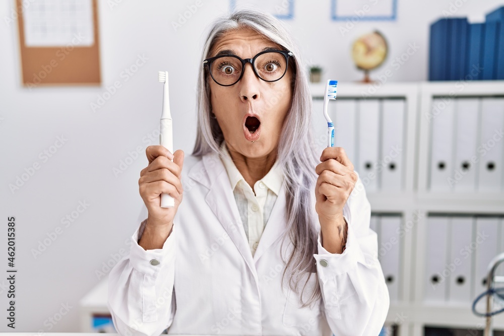Middle age grey-haired woman working at dentist clinic holding electric teethbrush and toothbrush in shock face, looking skeptical and sarcastic, surprised with open mouth Middle age grey-haired woman working at dentist clinic holding electric teethbrush and toothbrush in shock face, looking skeptical and sarcastic, surprised with open mouth