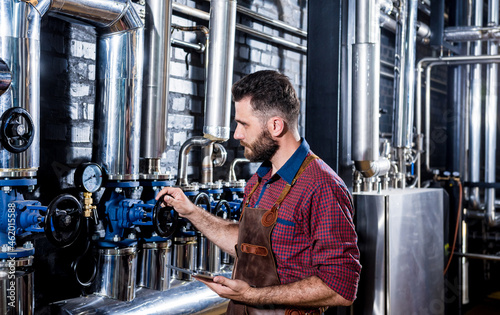 Young male brewer in leather apron supervising the process of beer fermentation at modern brewery factory