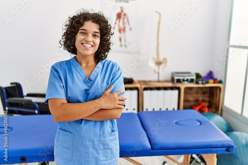 Young hispanic physiotherapist woman working at pain recovery clinic happy face smiling with crossed arms looking at the camera. positive person. photo