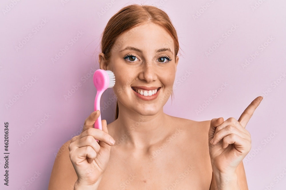 Young irish woman using facial exfoliating brush smiling happy pointing with hand and finger to the side Young irish woman using facial exfoliating brush smiling happy pointing with hand and finger to the side