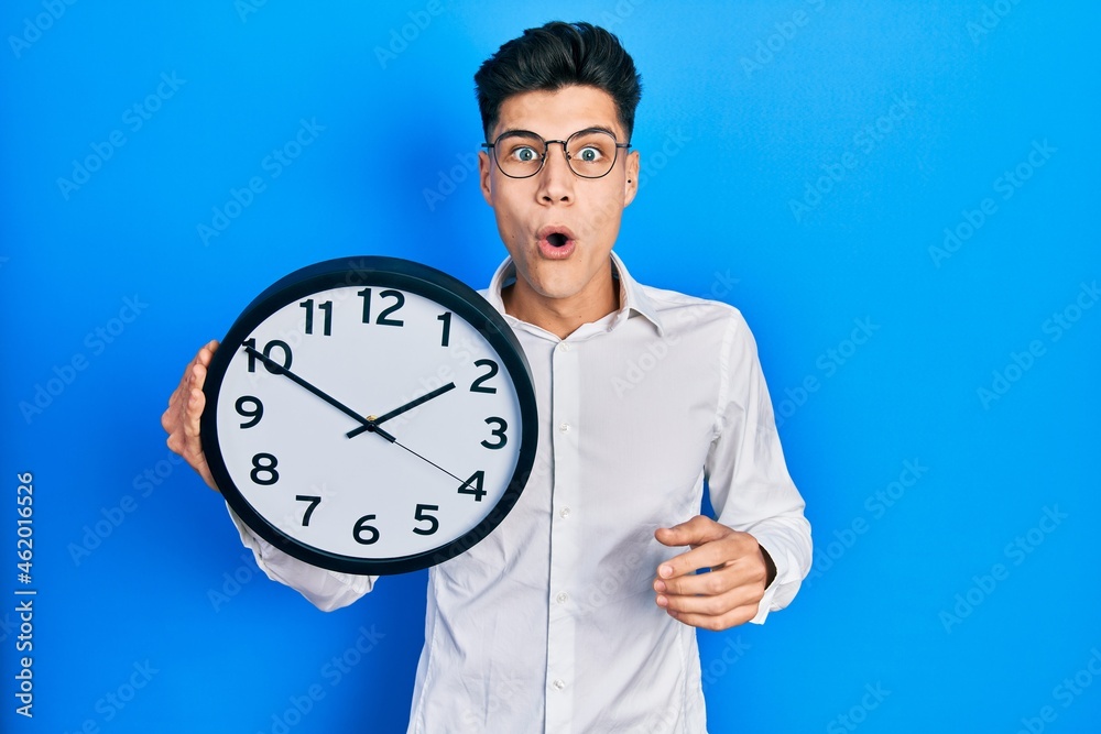 Young hispanic man holding big clock scared and amazed with open mouth for surprise, disbelief face Young hispanic man holding big clock scared and amazed with open mouth for surprise, disbelief face