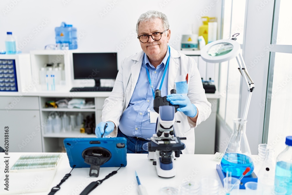 Middle age grey-haired man wearing scientist uniform using microscope and touchpad at laboratory Middle age grey-haired man wearing scientist uniform using microscope and touchpad at laboratory