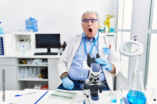 Senior caucasian man working at scientist laboratory in shock face, looking skeptical and sarcastic, surprised with open mouth