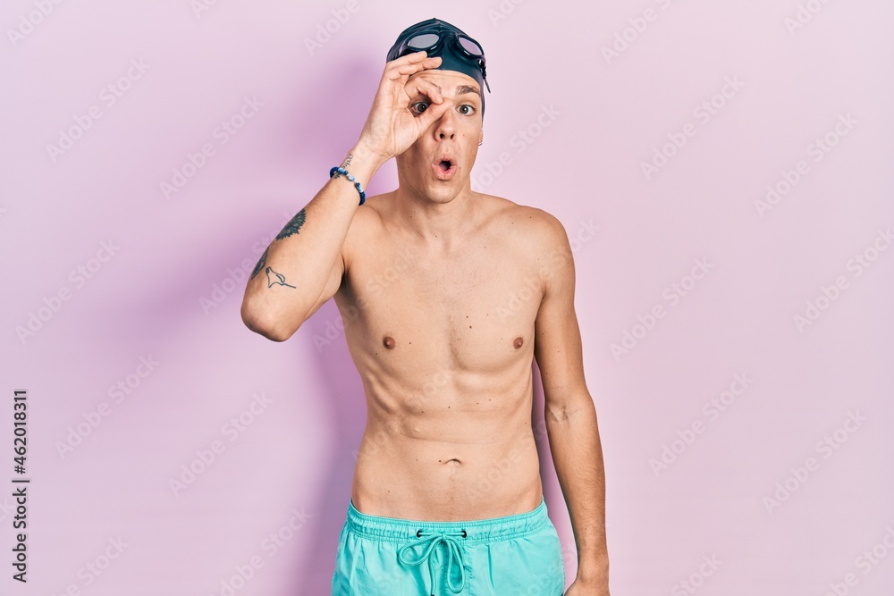 Young hispanic man wearing swimwear and swimmer glasses doing ok gesture shocked with surprised face, eye looking through fingers. unbelieving expression.