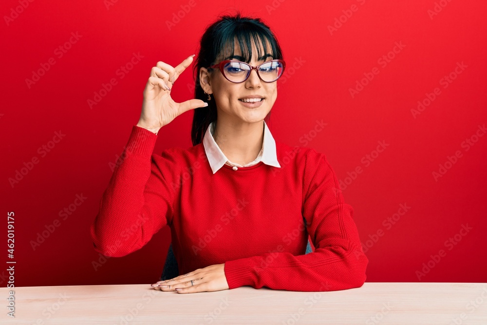 Young brunette woman with bangs wearing glasses sitting on the table smiling and confident gesturing with hand doing small size sign with fingers looking and the camera. measure concept. Young brunette woman with bangs wearing glasses sitting on the table smiling and confident gesturing with hand doing small size sign with fingers looking and the camera. measure concept.