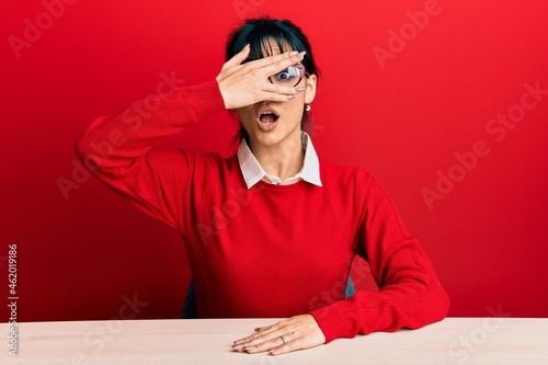 Young brunette woman with bangs wearing glasses sitting on the table peeking in shock covering face and eyes with hand, looking through fingers with embarrassed expression.