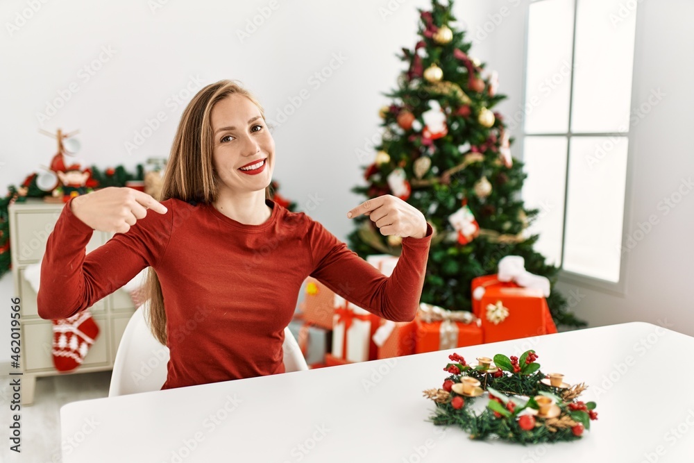 Caucasian young blonde woman sitting on the table by christmas tree looking confident with smile on face, pointing oneself with fingers proud and happy. Caucasian young blonde woman sitting on the table by christmas tree looking confident with smile on face, pointing oneself with fingers proud and happy.