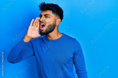 Arab man with beard wearing casual blue sweater shouting and screaming loud to side with hand on mouth. communication concept.