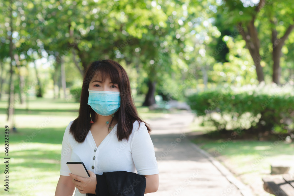 Fototapeta premium attractive business woman wearing face mask hold smartphone standing in park with sunrise and blur nature background. business new normal with corona virus or covid-19.